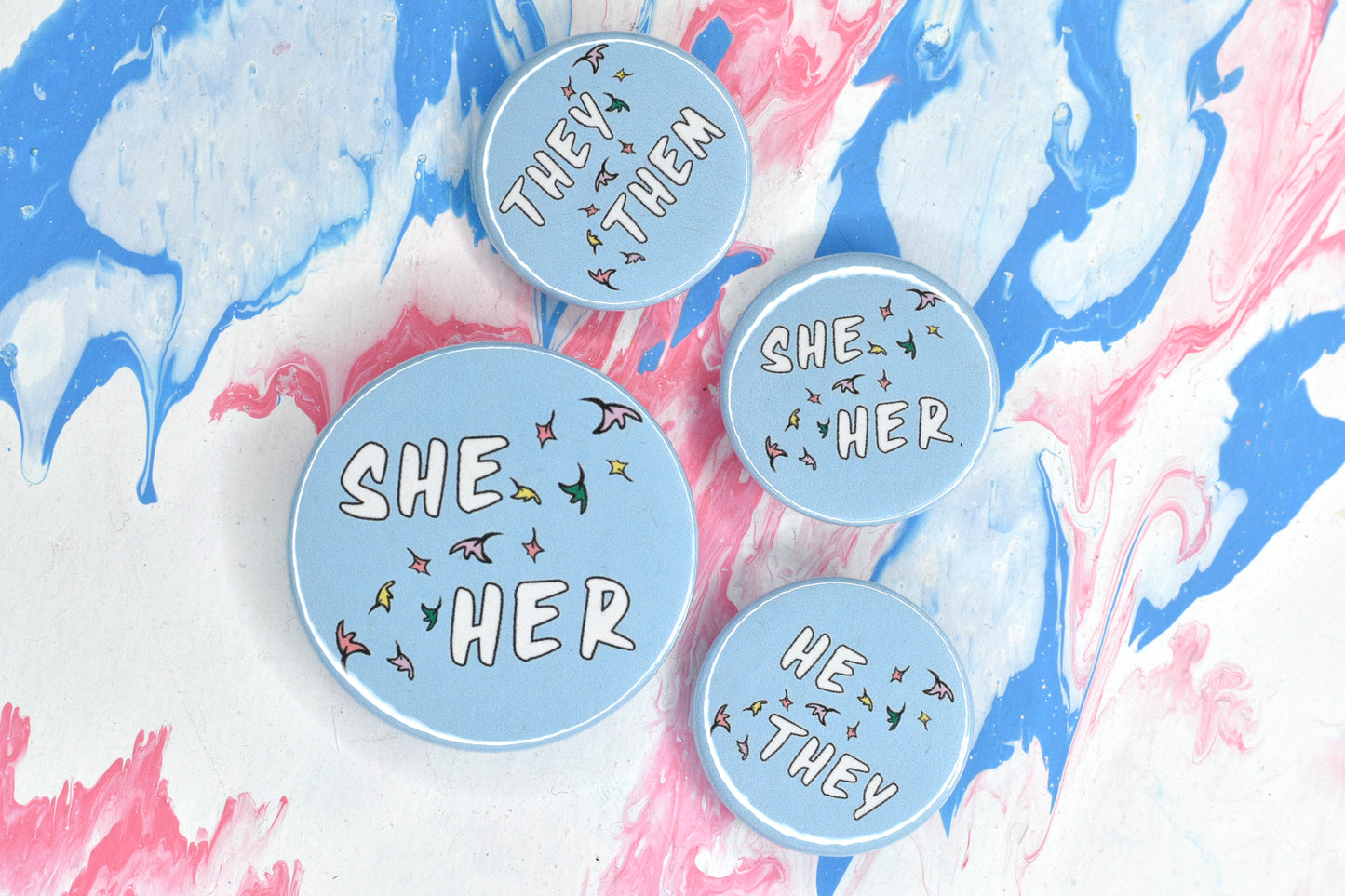 A photo of four round pin badges. One is size medium and the other three are size small. Each has a pastel blue background and a set of pronouns in uppercase white text. The pronouns are separated by a diagonal band of pastel coloured comic book style leaves. The pronouns on the medium badge are "she her". The pronouns on the small badges are "they them", "she her" and "he they".