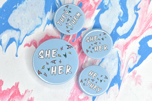 A photo of four round pin badges. One is size medium and the other three are size small. Each has a pastel blue background and a set of pronouns in uppercase white text. The pronouns are separated by a diagonal band of pastel coloured comic book style leaves. The pronouns on the medium badge are "she her". The pronouns on the small badges are "they them", "she her" and "he they".