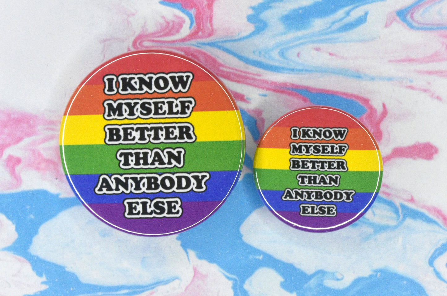 Two circular pin badges in sizes medium and small each with a rainbow pride flag background and black and white text in a bubble-like font reading "I know myself better than anybody else".