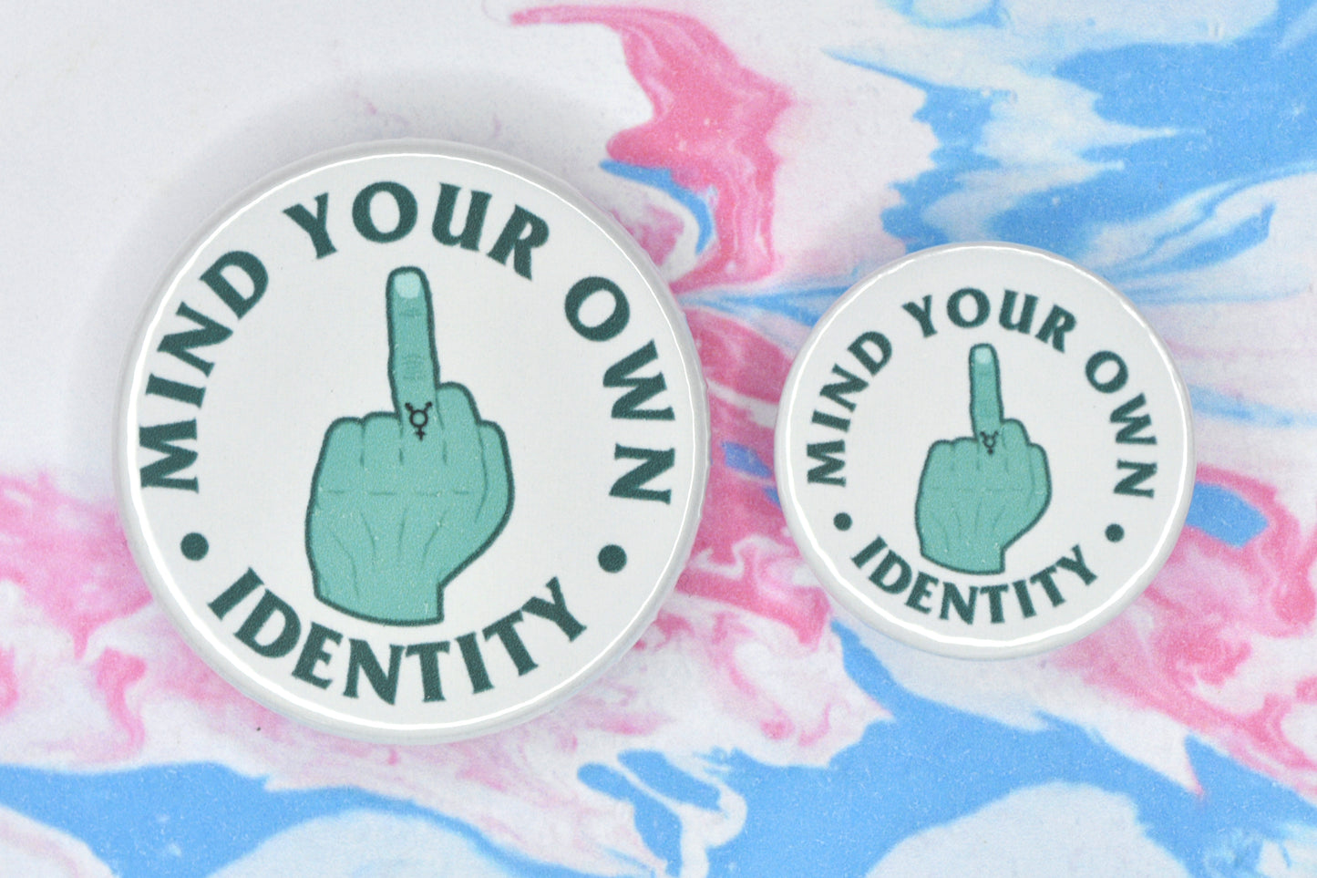 Two badges in sizes medium and small respectively, each featuring a white background with an illustration of a hand giving the middle finger in the centre. The hand is teal in colour and has a trans symbol tattoo on the middle finger. There is text around the illustration in a darker teal shade which reads "mind your own identity".