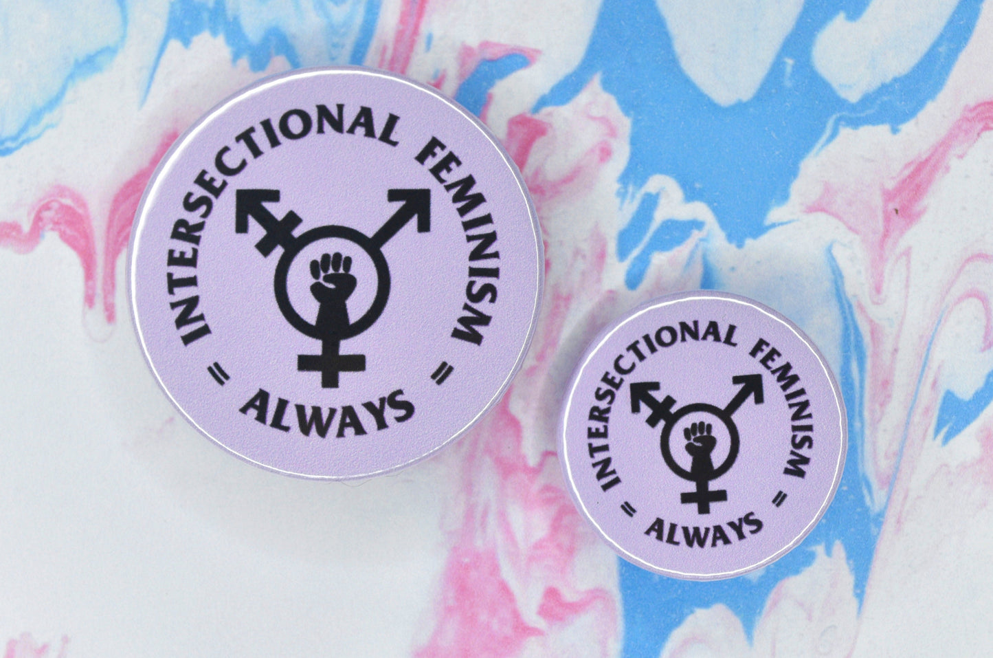 Two circular pin badges in sizes medium and small each with a light purple background and an intersectional feminism logo in the centre, i.e. a trans symbol with a fist in the centre. There is text in a circle surrounding this logo which reads "intersectional feminism always".