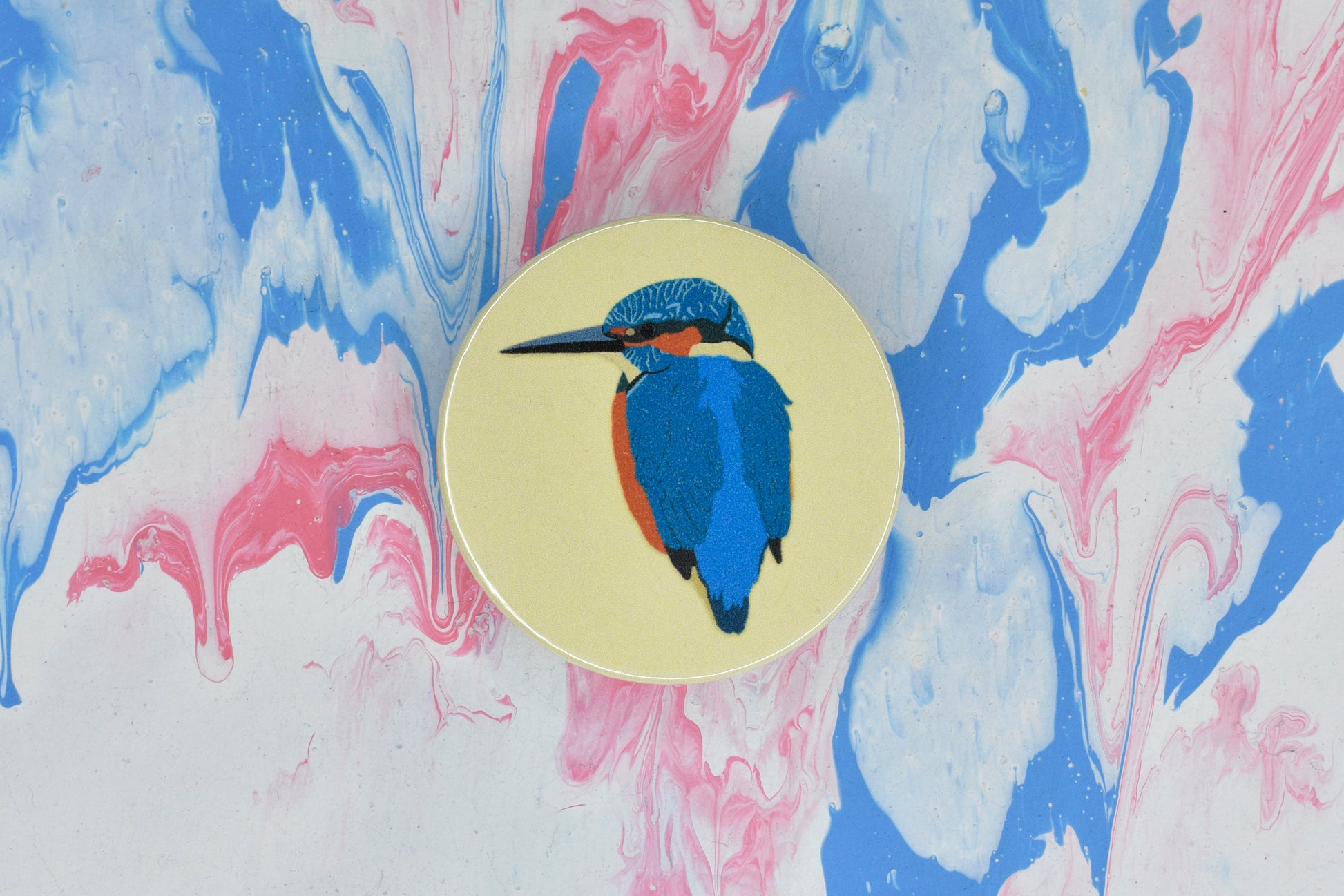 A circular pin badge with a pastel yellow background featuring an illustration of a kingfisher, a blue bird with orange on its chest and around its face.