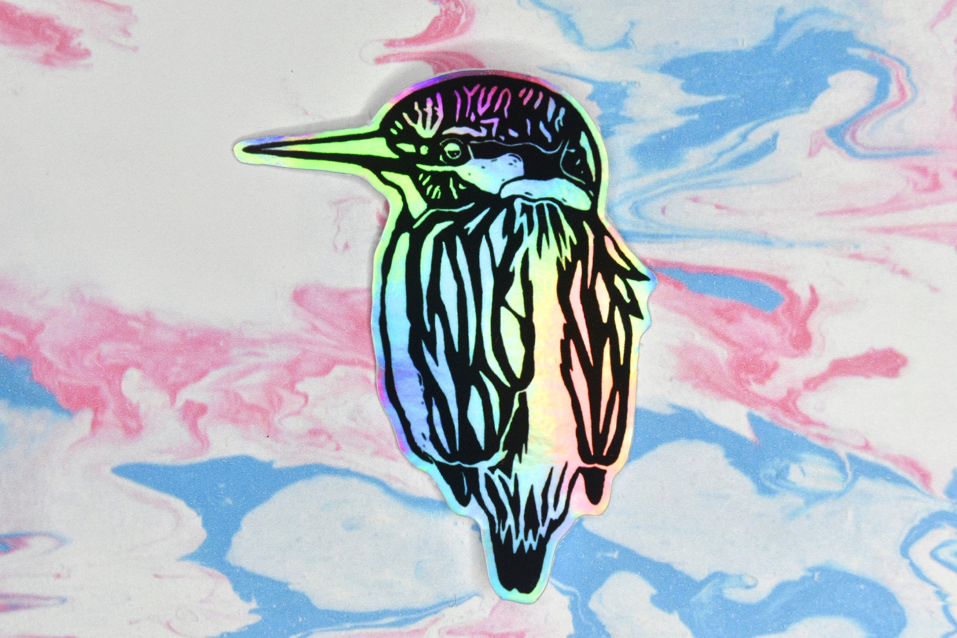 A sticker featuring a kingfisher illustration in black ink on an iridescent holographic background. The holographic effect produces a metallic rainbow of colour across the surface of the sticker.