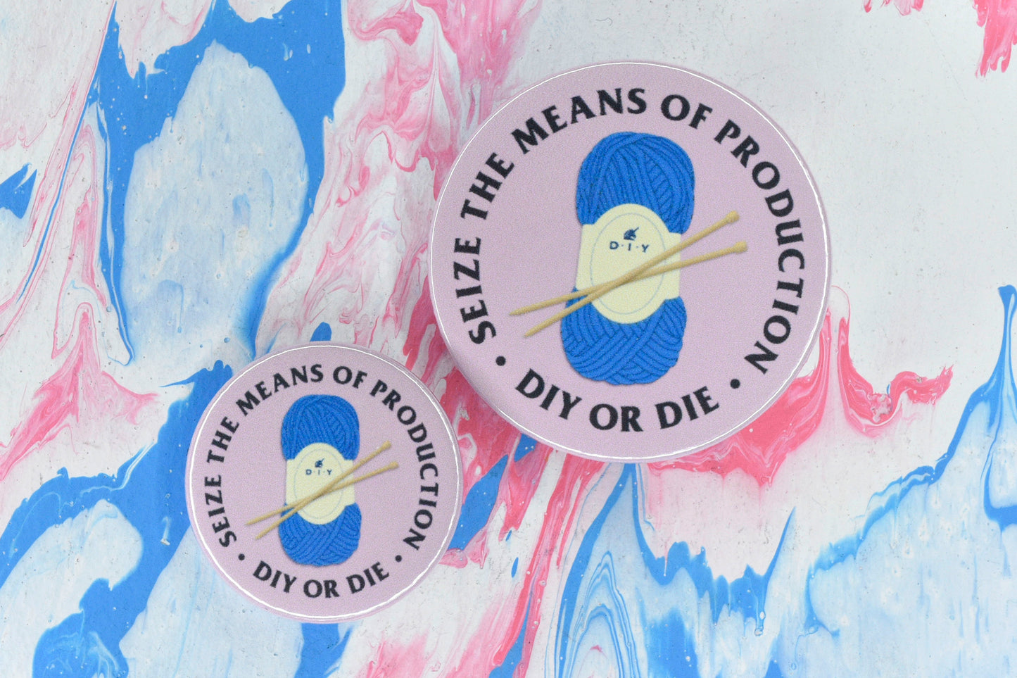 Two circular pin badges in sizes medium and small, each with a light pink background and an illustration of a ball of blue yarn and two wooden knitting needles. There is uppercase black serif text surrounding this reading "seize the means of production - DIY or die".