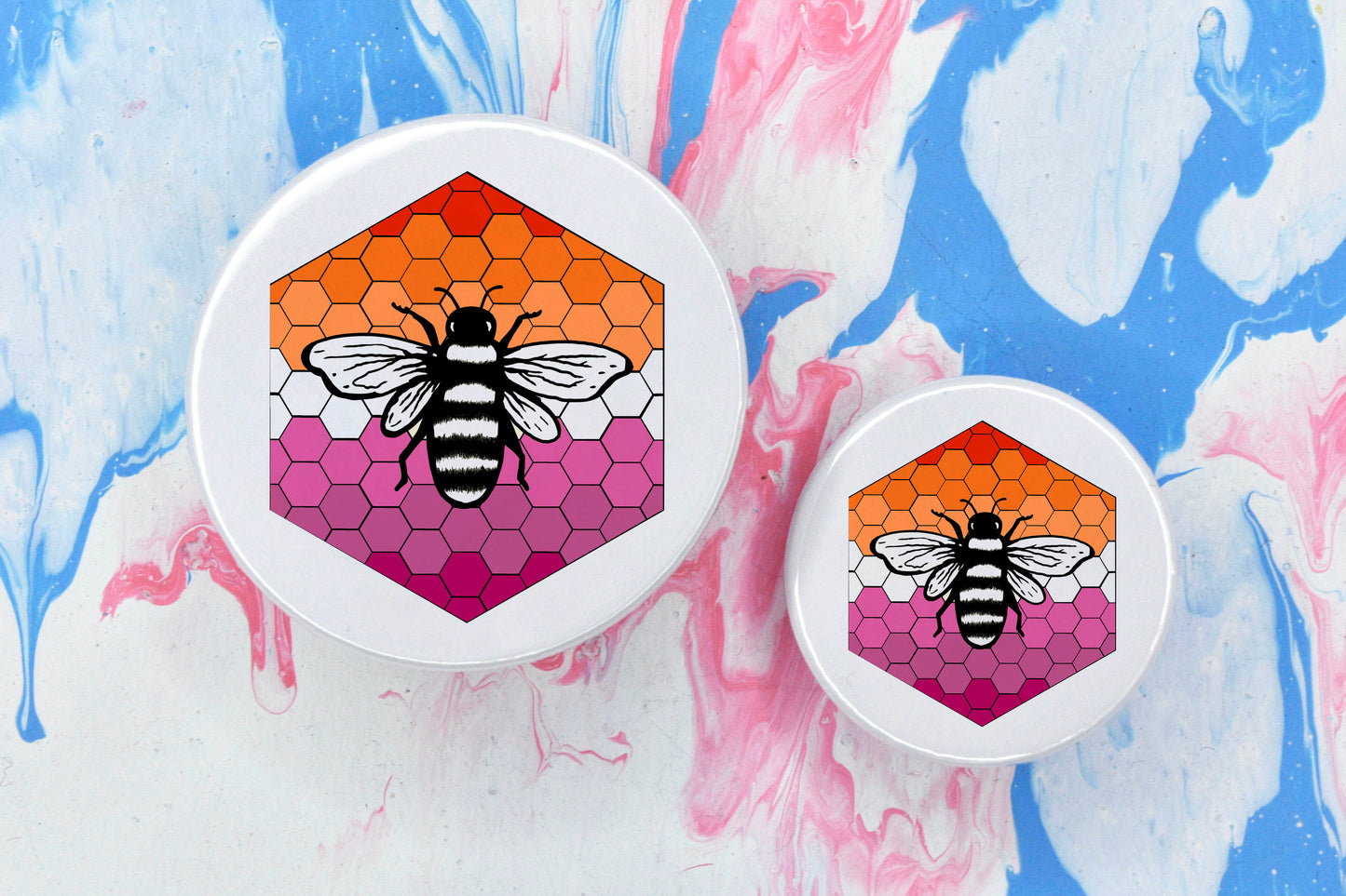 Two badges in sizes medium and small respectively, each featuring a white background with a black and white illustration of a bee overlaid on a bee hive pattern which is coloured in with the lesbian pride flag colours (orange, white and pink).