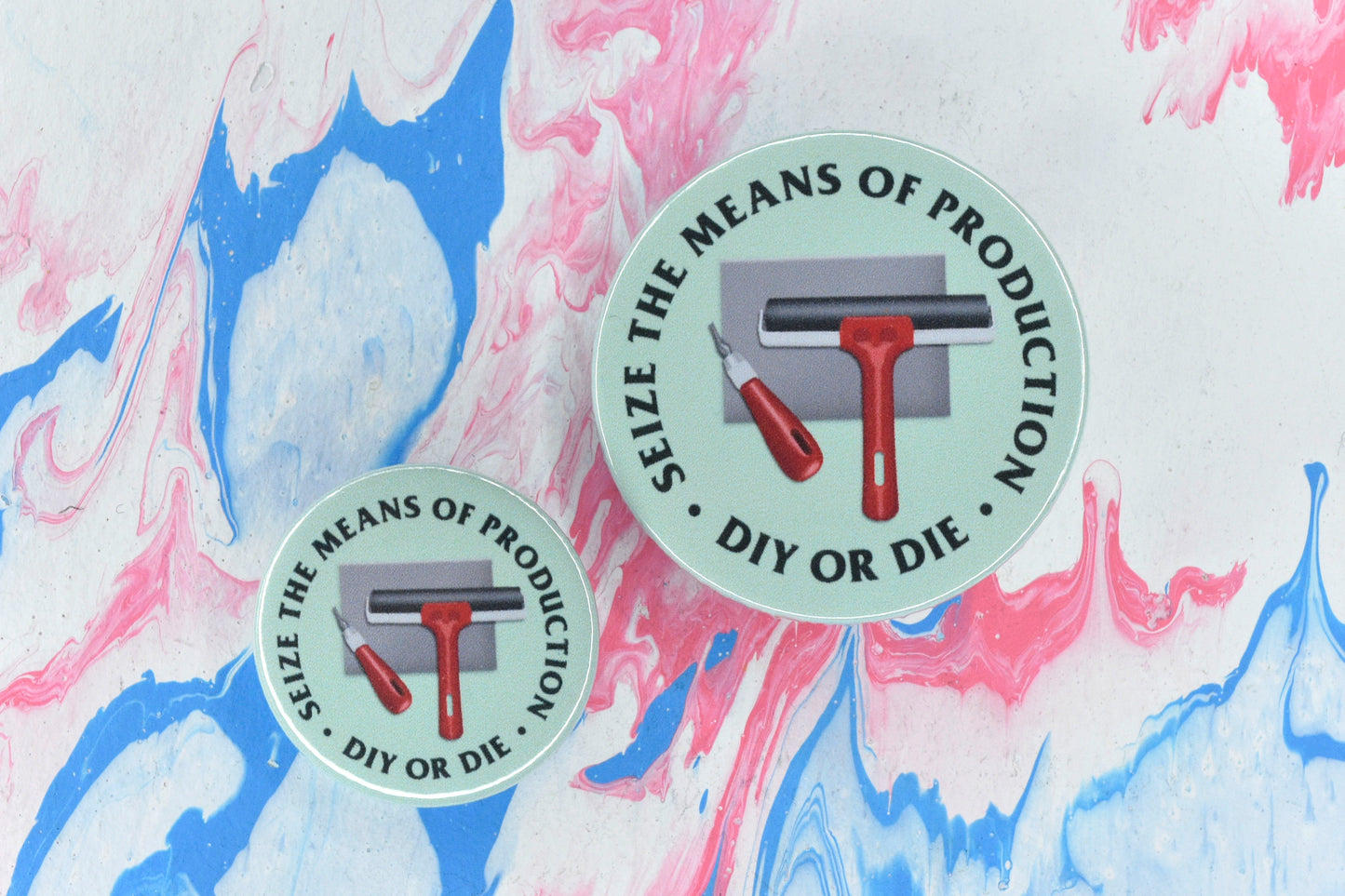 Two circular pin badges in sizes medium and small, each with a light green background and an illustration of a red handled lino cutting tool and lino roller on a grey lino block. There is uppercase black serif text surrounding this reading "seize the means of production - DIY or die".