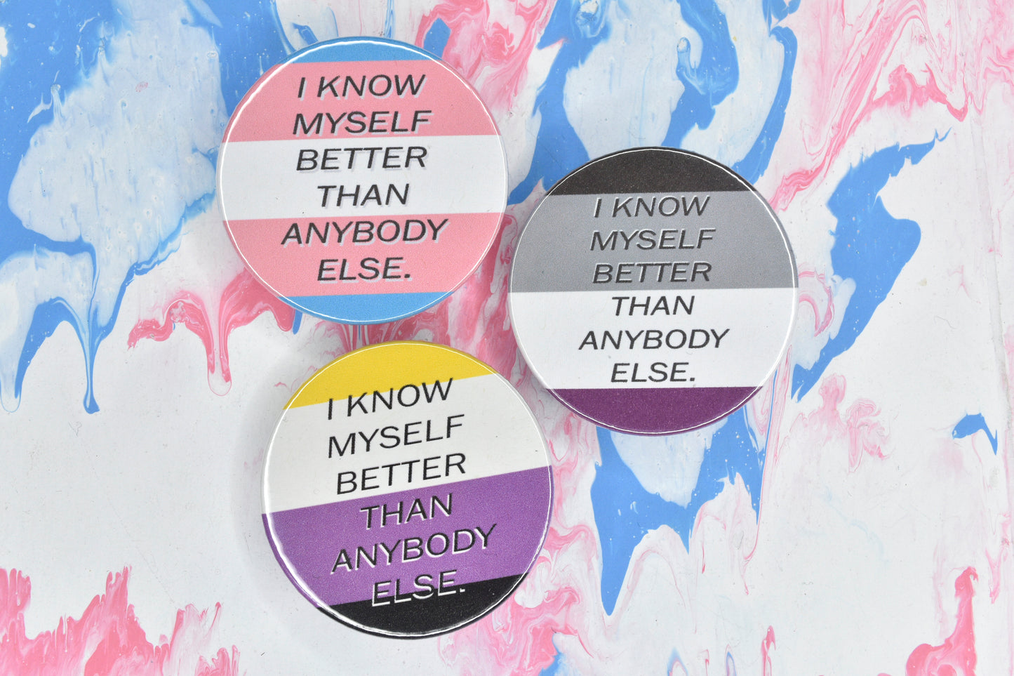 Three pin badges, each featuring a different pride flag with black uppercase sans serif text overlaid reading "I know myself better than anybody else." The pride flags shown are trans, asexual and non-binary.