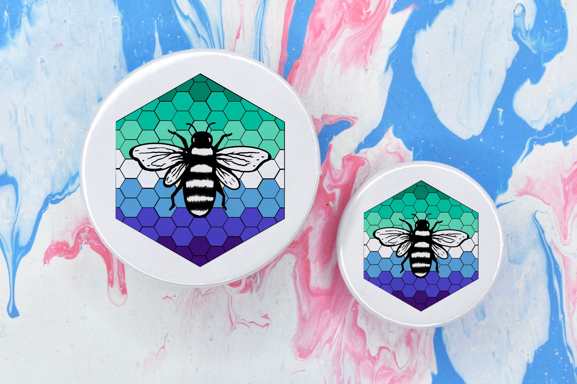 Two badges in sizes medium and small respectively, each featuring a white background with a black and white illustration of a bee overlaid on a bee hive pattern which is coloured in with the mlm pride flag colours (greens, white and blues).