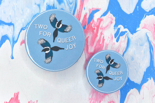 Two pin badges, one in size medium and the other in size small, both with a light blue background and featuring an illustration of two flying magpies with white text curving around them which reads "two for queer joy".