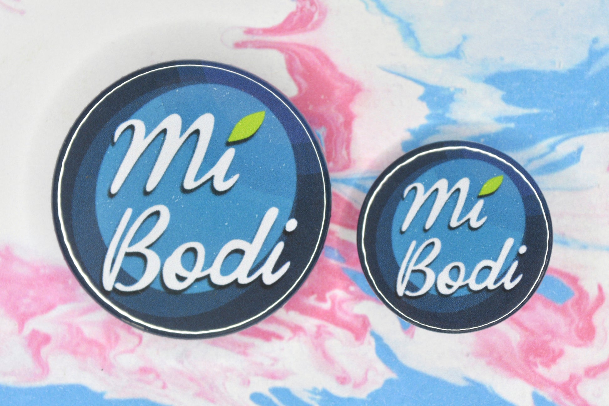  Two badges in sizes medium and small respectively, each featuring a background made up of different blue shades and white text reading "mi bodi" with a green leaf over the letter I in "Mi"