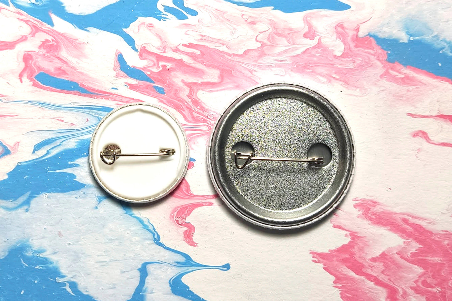 A photograph showing the back of two badges, in sizes small and medium respectively. The small badge features a white plastic backing with a metal safety pin closure. The medium badge features a silver metal backing with a safety pin closure.