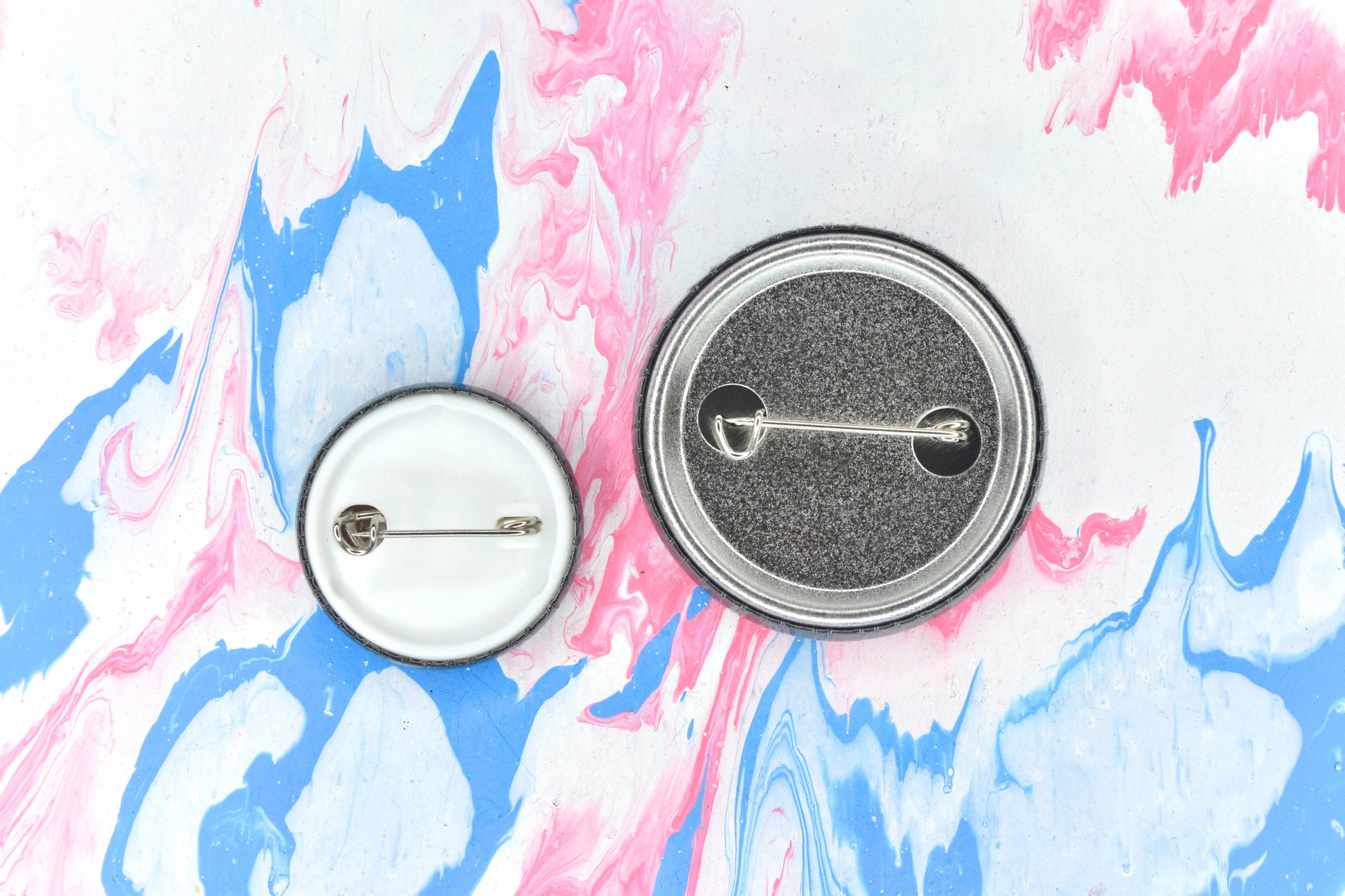 A photograph showing the back of two badges, in sizes small and medium respectively. The small badge features a white plastic backing with a metal safety pin closure. The medium badge features a silver metal backing with a safety pin closure.