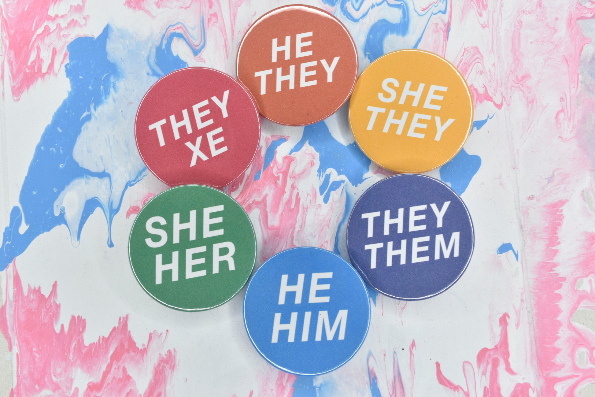 Six pronoun pins, each with a solid colour background and white text featuring various sets of pronouns. The pins are shown in dark pink with pronouns "they/xe", orange with pronouns "he/they", yellow with pronouns "she/they", purple with "they/them", blue with "he/him" and green with "she/her".