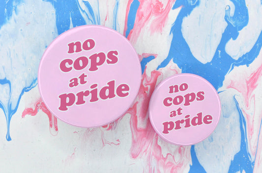 Two circular pin badges in sizes medium and small each with a light pink background and dark pink text in a bubble-like font reading "no cops at pride".