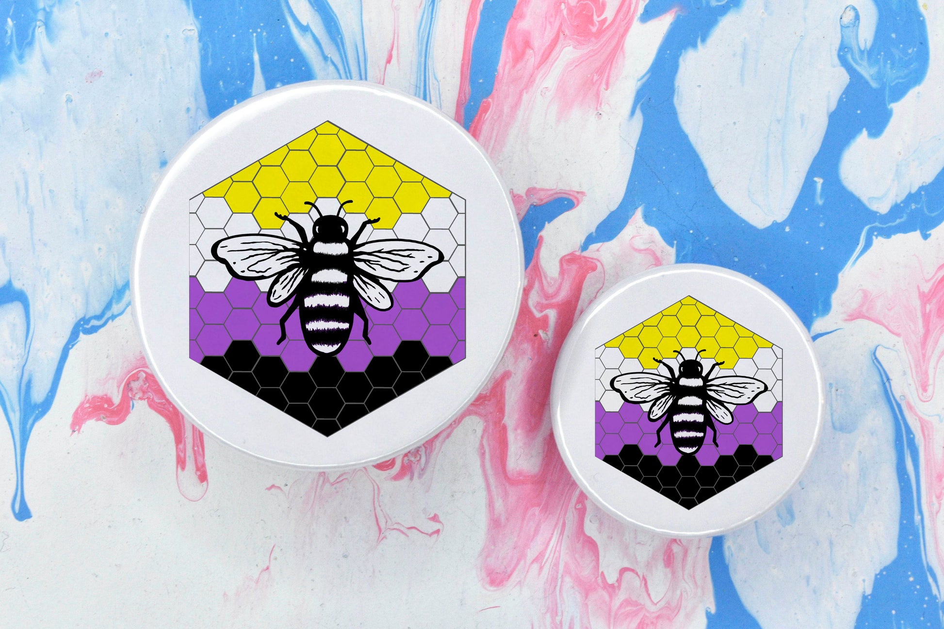 Two badges in sizes medium and small respectively, each featuring a white background with a black and white illustration of a bee overlaid on a bee hive pattern which is coloured in with the non-binary pride flag colours (yellow, white, purple and black).