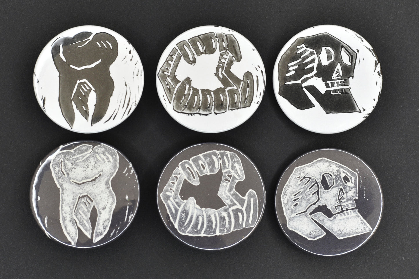 A group of six badges, each featuring either a black background with an original lino print stamped design in white ink, or a white background with an original lino print stamped design in black ink. The designs are a skull, a set of fangs and a single tooth.