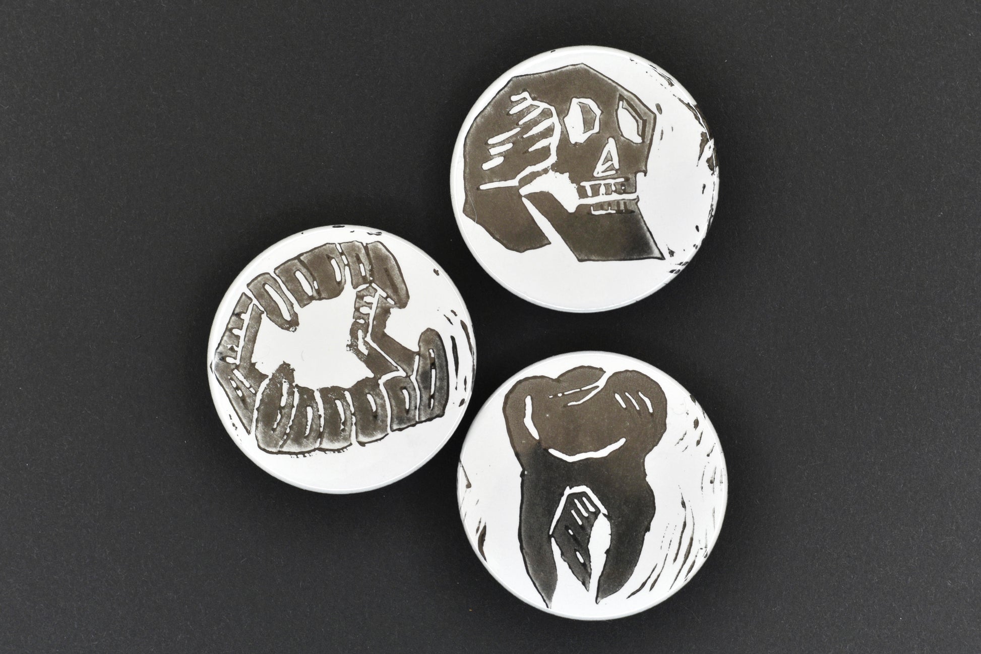 A group of three badges, each featuring a white background with an original lino print stamped design in black ink. The designs are a skull, a set of fangs and a single tooth.