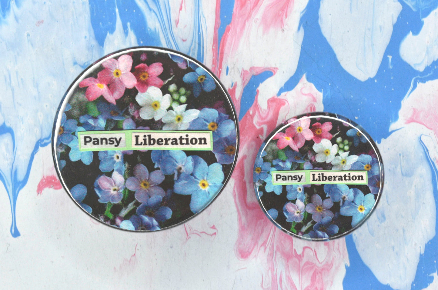 Two pin badges in sizes medium and small, both featuring a collage consisting of a photo of pink, white and blue forget-me-not flowers with newspaper print overlaid reading "pansy liberation".