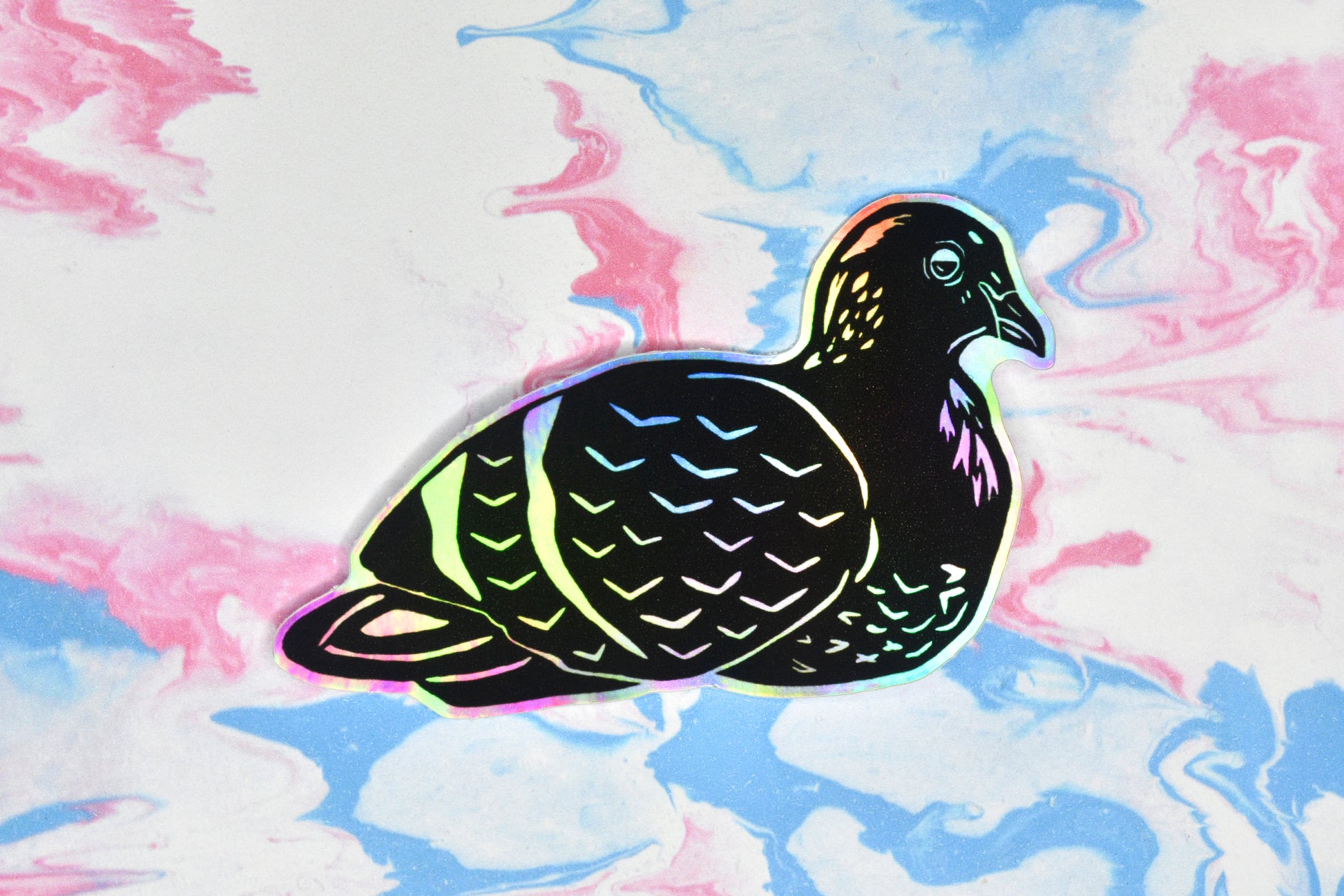 A sticker featuring a pigeon illustration in black ink on an iridescent holographic background. The holographic effect produces a metallic rainbow of colour across the surface of the sticker.