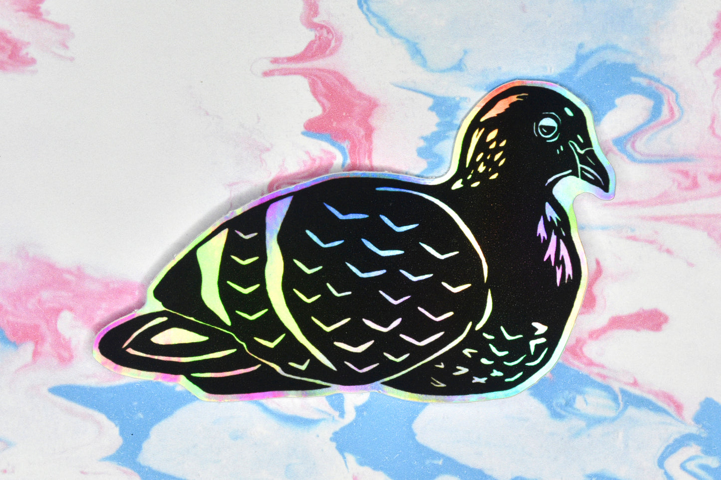 A sticker featuring a pigeon illustration in black ink on an iridescent holographic background. The holographic effect produces a metallic rainbow of colour across the surface of the sticker.