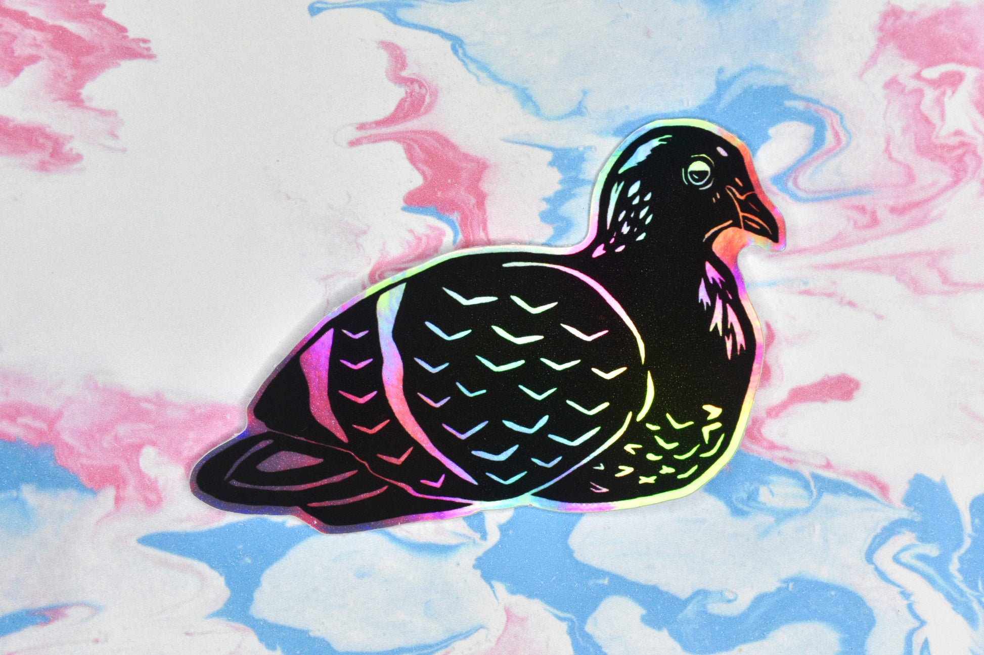 A sticker featuring a pigeon illustration in black ink on an iridescent holographic background. The holographic effect produces a metallic rainbow of colour across the surface of the sticker.