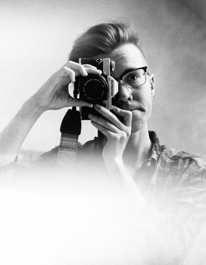 A black and white self-portrait of Alli, a white man in his 20s with dark brown hair and glasses. He is holding an analogue camera to his eye and is taking the photo in a mirror