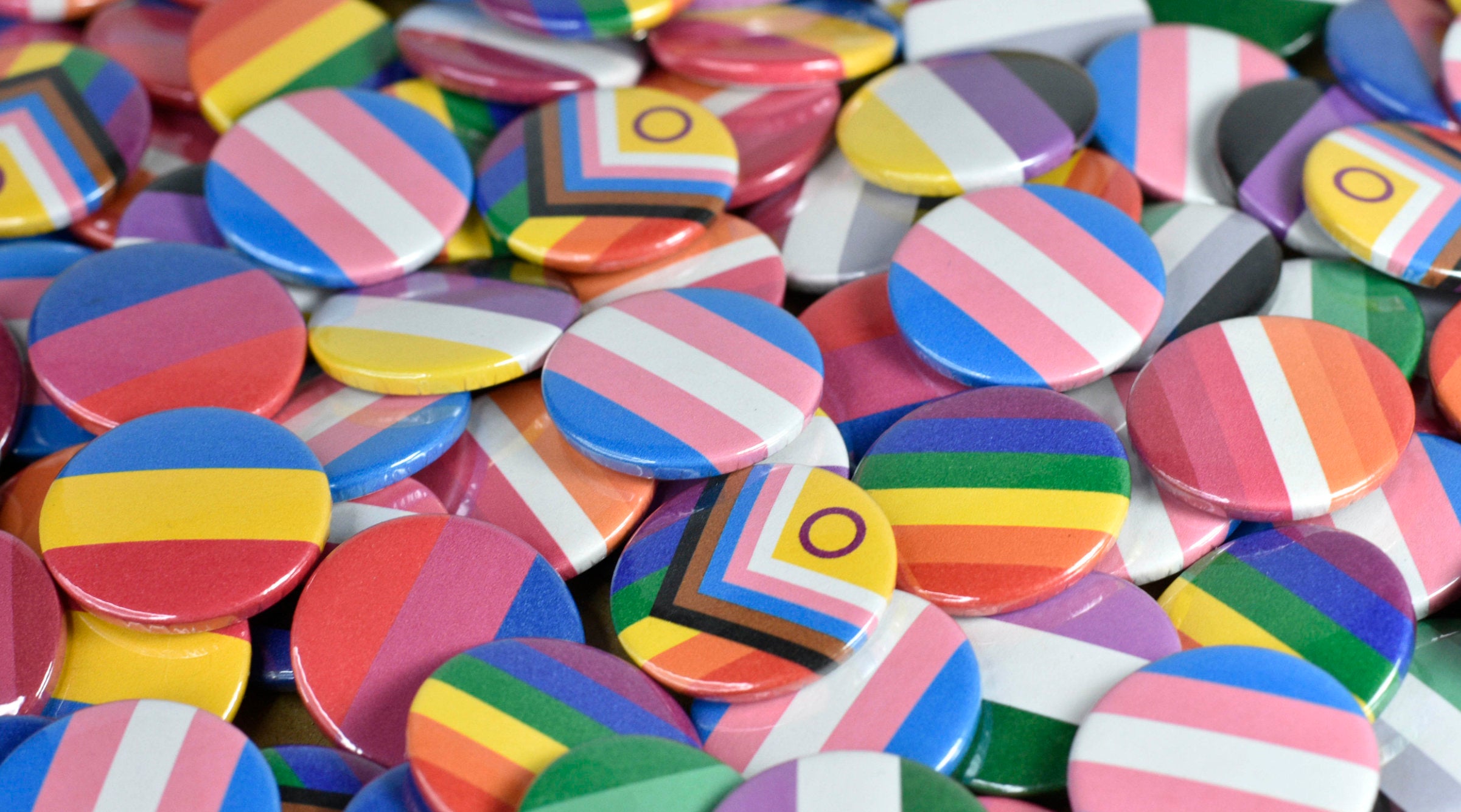 A large pile of small circular pin badges featuring a range of pride flags including progress pride, rainbow, trans, non-binary, asexual, bisexual, pansexual, lesbian and aromantic.