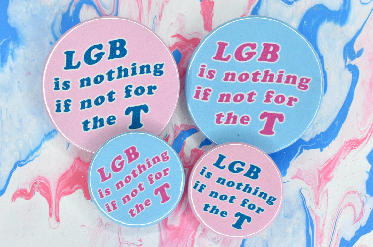 A photograph showing four pin badges, each with a variation of the same design. There is a medium and a small pin, both with a light blue background and large pink text reading "LGB is nothing if not for the T". There is another medium and small pin, both with a light pink background and large blue text reading "LGB is nothing if not for the T".