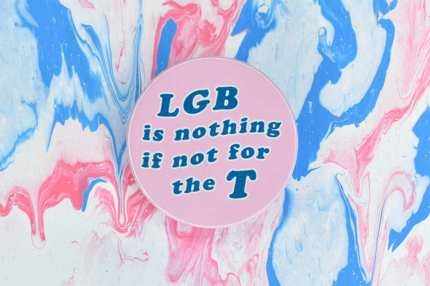 A photograph showing a pin badge with a light pink background and large blue text reading "LGB is nothing if not for the T".