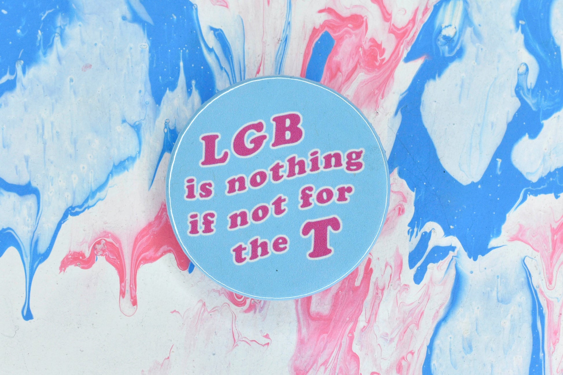 A photograph showing a pin badge with a light blue background and large pink text reading "LGB is nothing if not for the T".