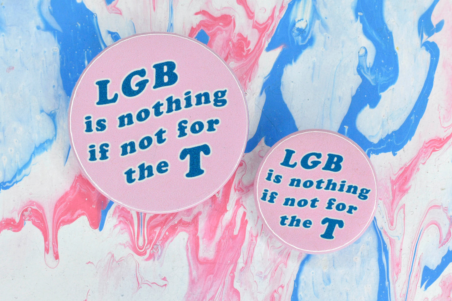 Two circular badges in sizes medium and small respectively, each with a light pink background and large blue text reading "LGB is nothing if not for the T".