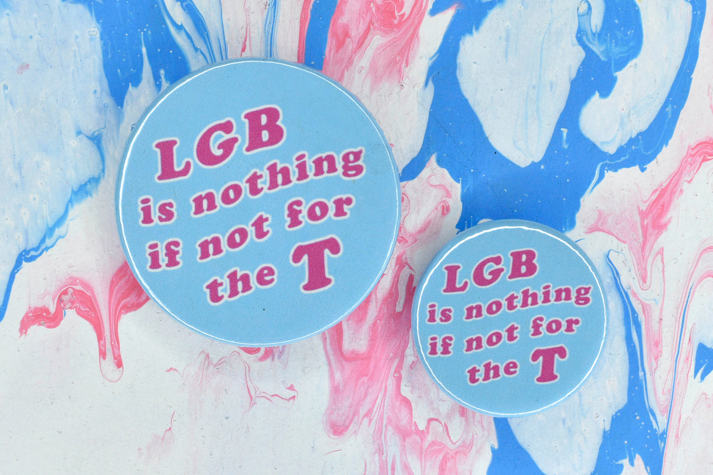 Two circular badges in sizes medium and small respectively, each with a light blue background and large pink text reading "LGB is nothing if not for the T".