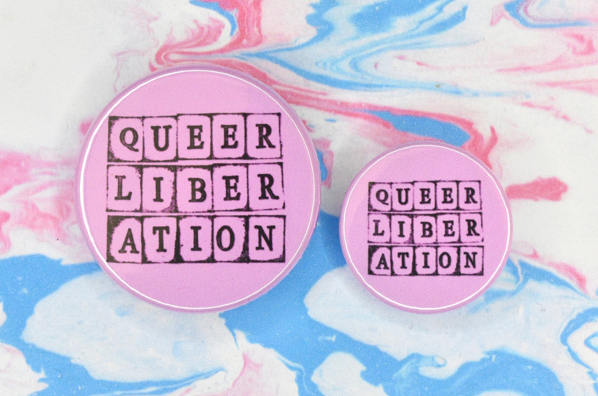 Two badges in sizes medium and small respectively, each featuring a pink background with text in a hand-stamped font reading "queer liberation".