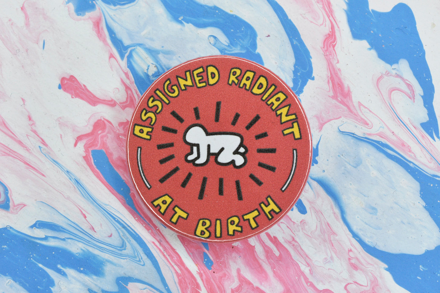 A pin badge with a red background and Keith Haring's radiant baby illustration in the centre in black and white. There is yellow text around the outside reading "assigned radiant at birth".