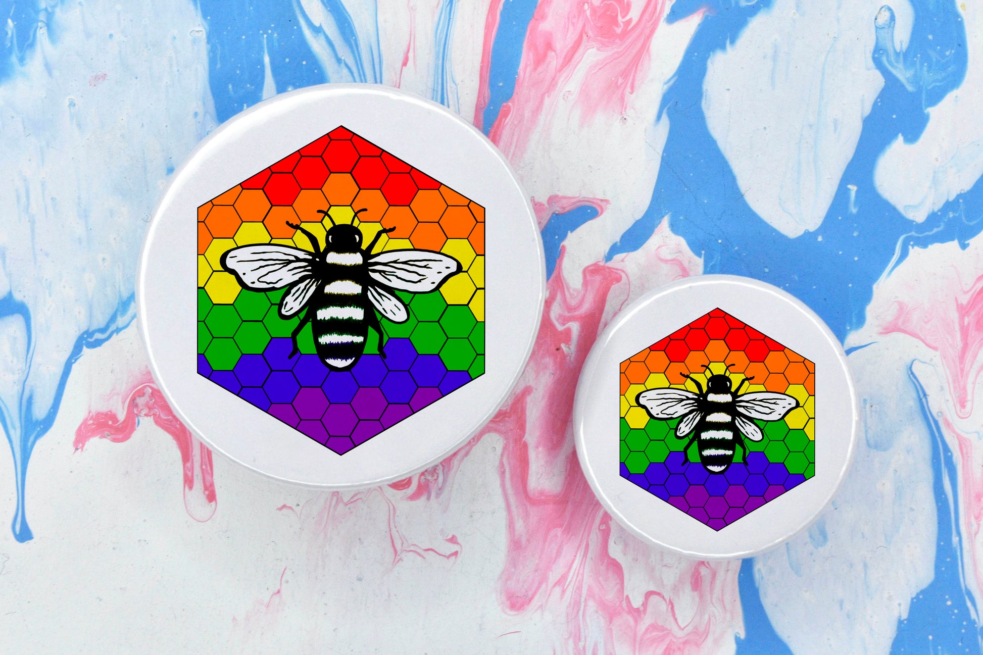 Two badges in sizes medium and small respectively, each featuring a white background with a black and white illustration of a bee overlaid on a bee hive pattern which is coloured in with the rainbow pride flag colours (red, orange, yellow, green, blue and purple).