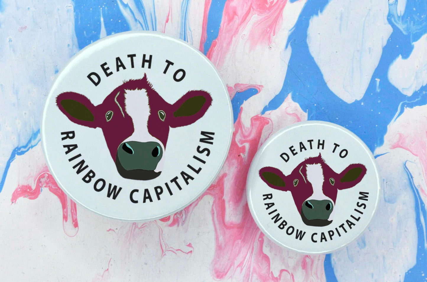 Two pin badges in sizes medium and small featuring an illustration of a cow with rainbow gradient text reading "death to rainbow capitalism" on a white background.