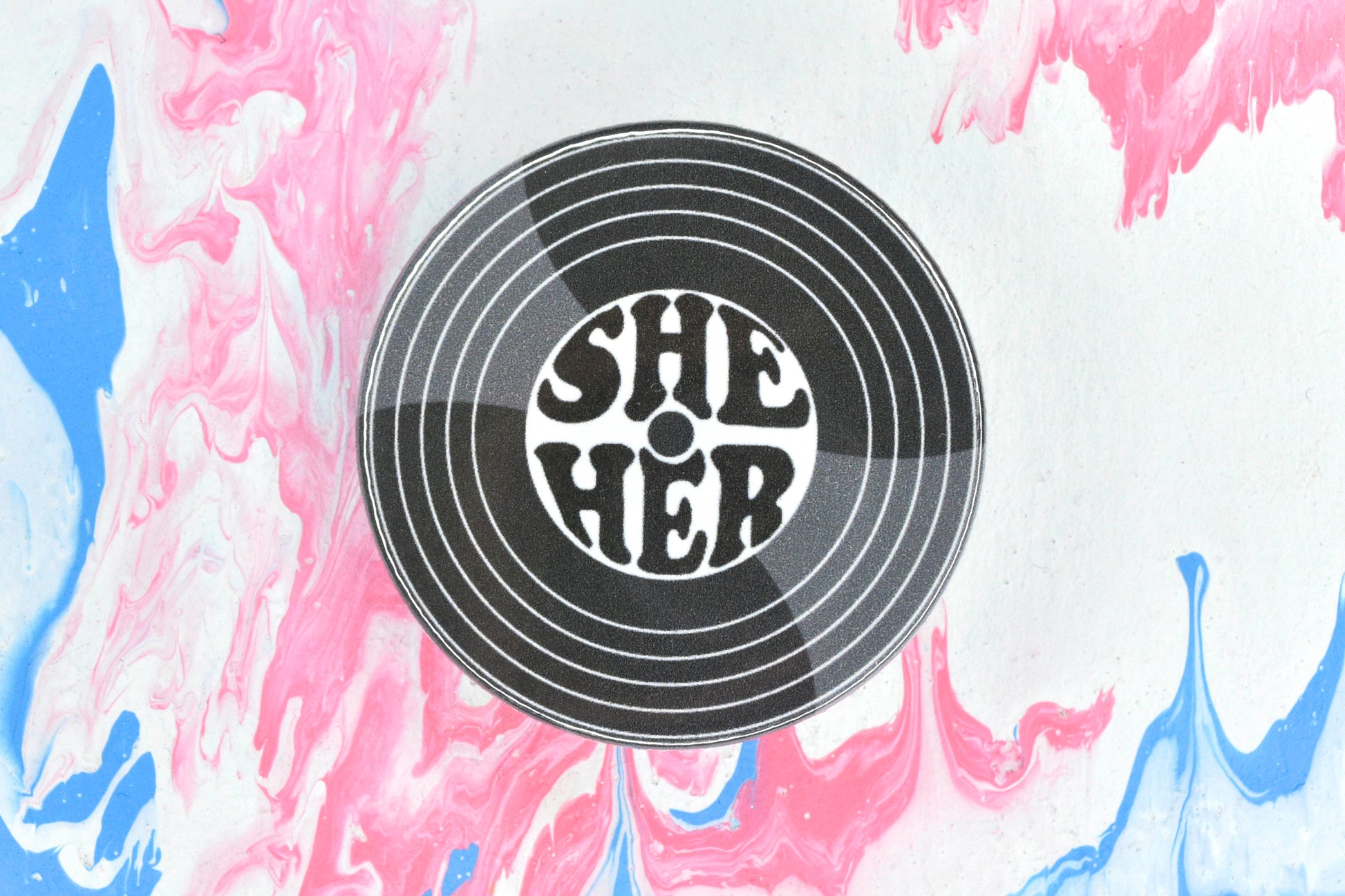 A circular badge, featuring an illustration on a vinyl record with a set of pronouns (she/her) in the centre.