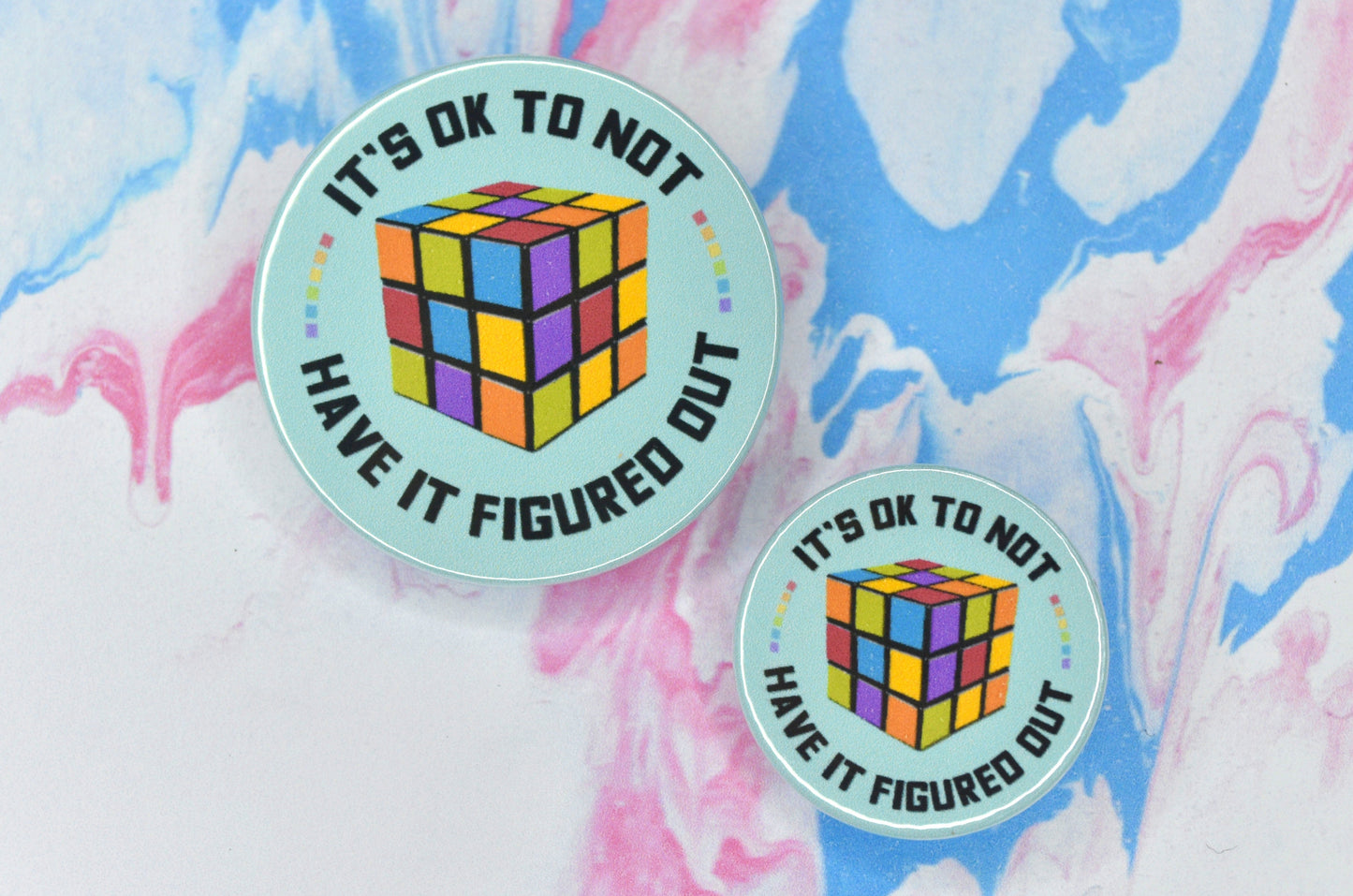 Two badges in sizes medium and small respectively, each featuring a mint green background with an illustration of a Rubik's cube in the centre. There is text in a circle around the illustration reading "it's ok to not have it figured out" with some small rainbow coloured squares separating the words.