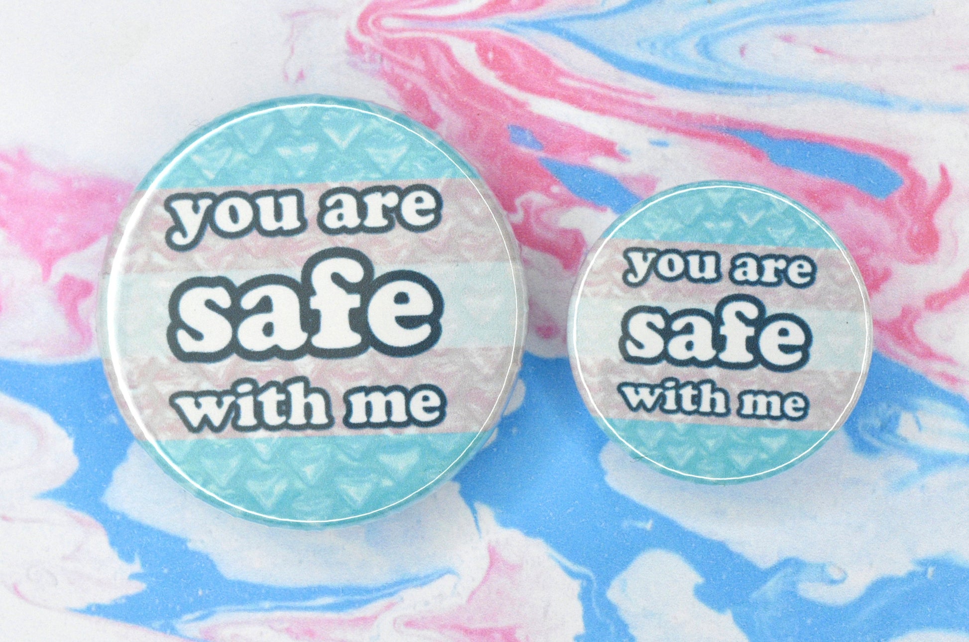 Two badges in sizes medium and small respectively, each featuring a trans flag background with heart-shaped bubble wrap overlaid. There is text on top of this in a bubble-like font reading "you are safe with me".