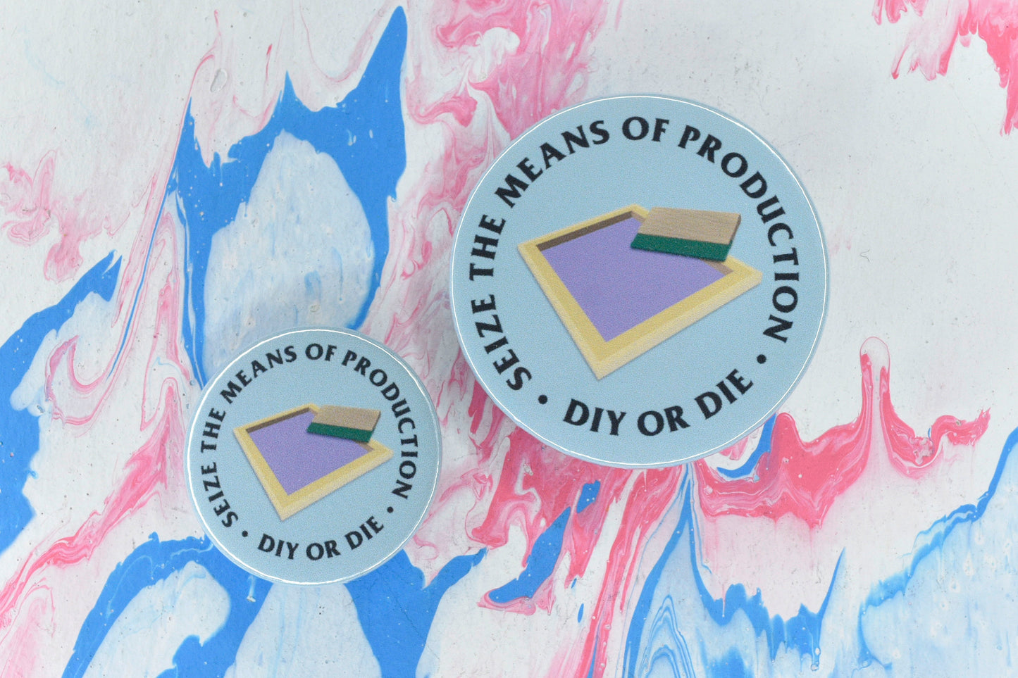 Two circular pin badges in sizes medium and small, each with a light blue background and an illustration of a screen printing screen and squeegee. There is uppercase black serif text surrounding this reading "seize the means of production - DIY or die".