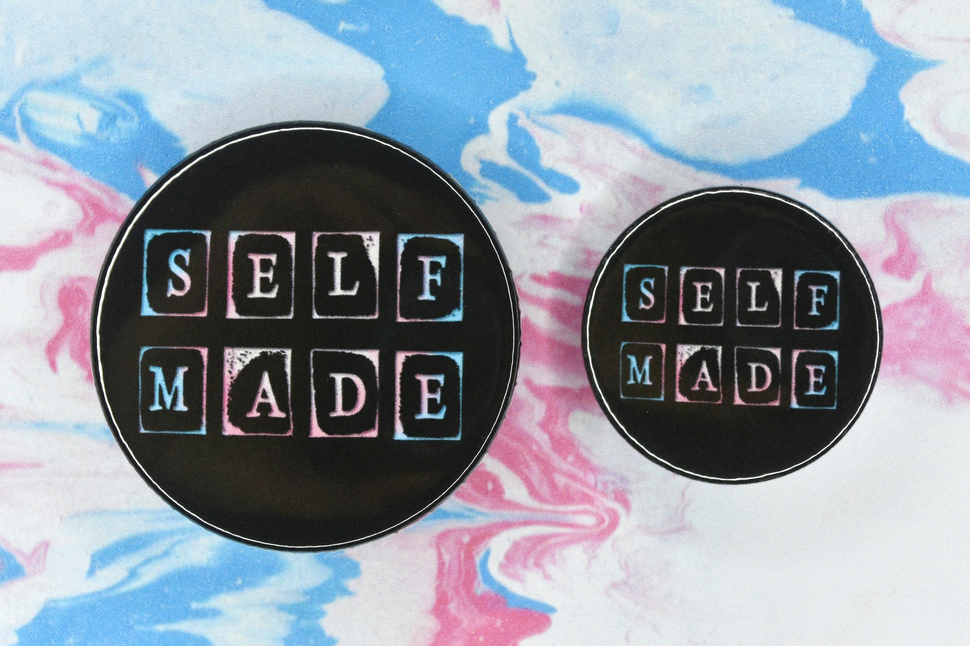 Button badges in two sizes featuring a trans pride design. Text in a hand-stamped font reads "Self Made". It has a trans pride flag colour effect against a black background.