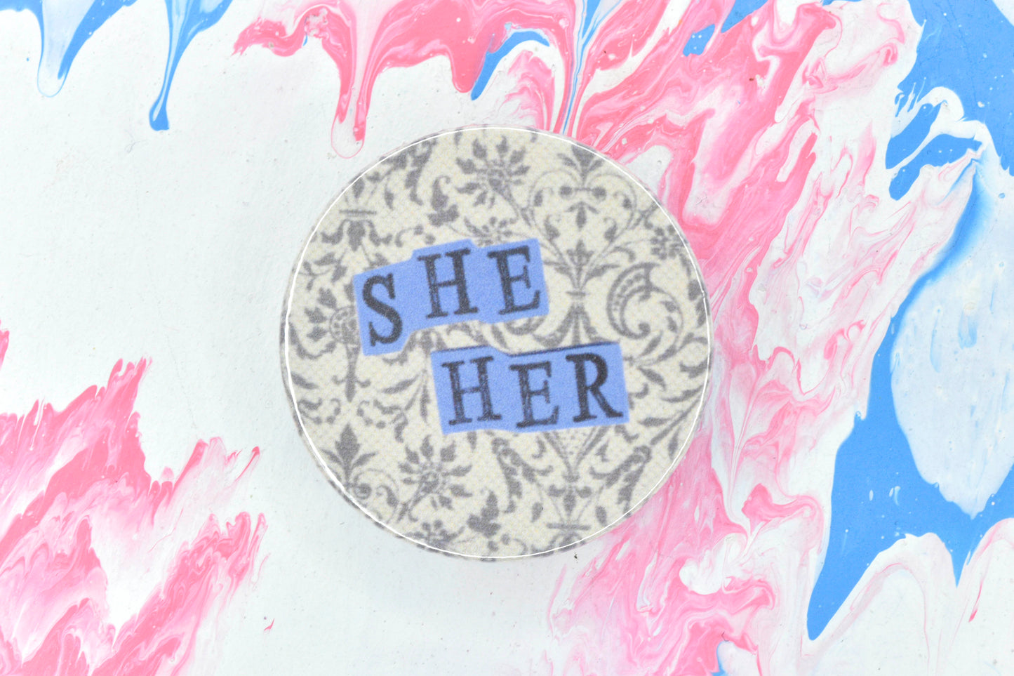 A pin badge featuring an off-white background with a lace pattern in dark grey. Overlaid are hand cut pieces of purple paper with hand stamped uppercase black lettering reading "she" and "her".