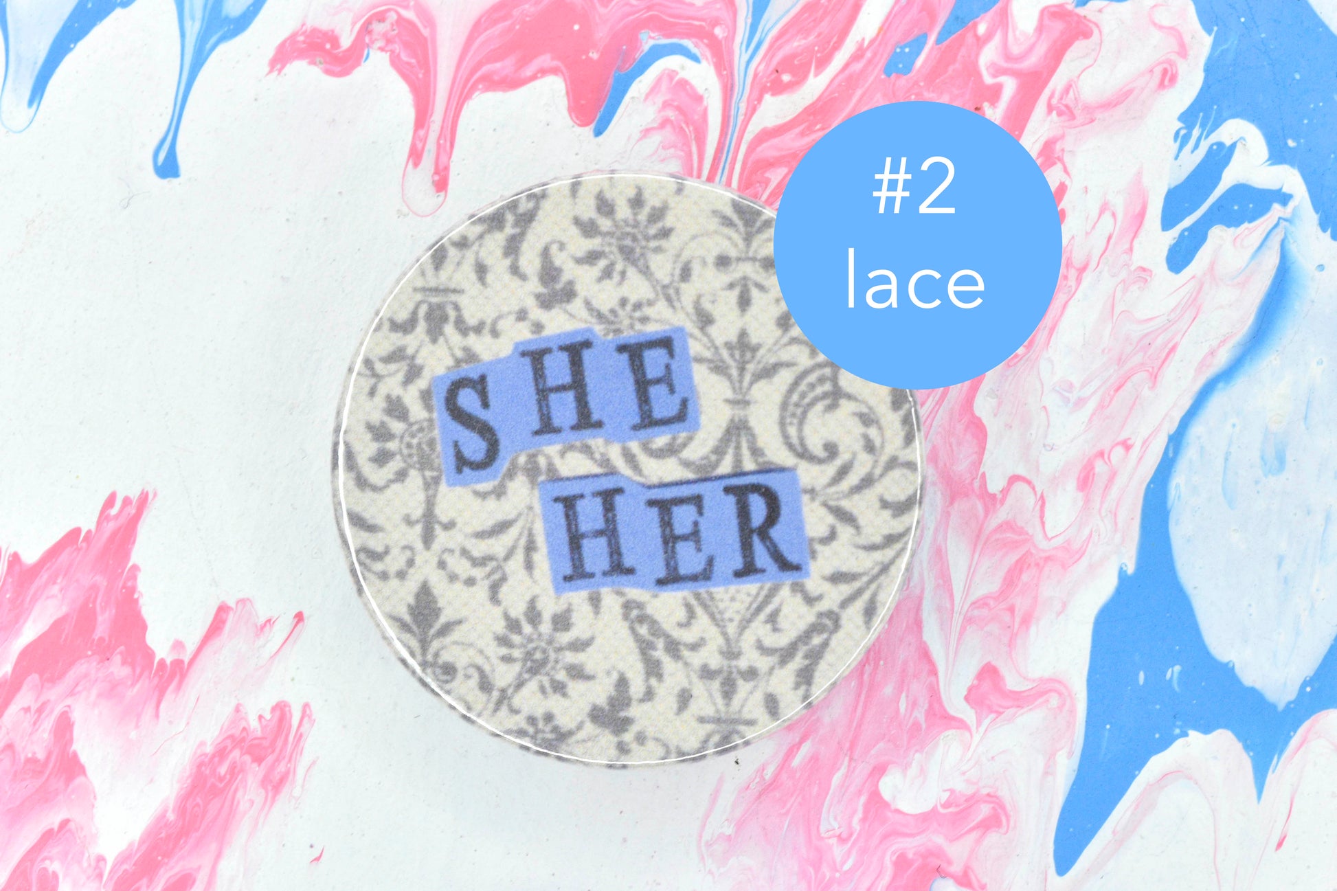 A pin badge featuring an off-white background with a lace pattern in dark grey. Overlaid are hand cut pieces of purple paper with hand stamped uppercase black lettering reading "she" and "her". There is a circular blue label next to the badge with white text reading "number 2, lace".