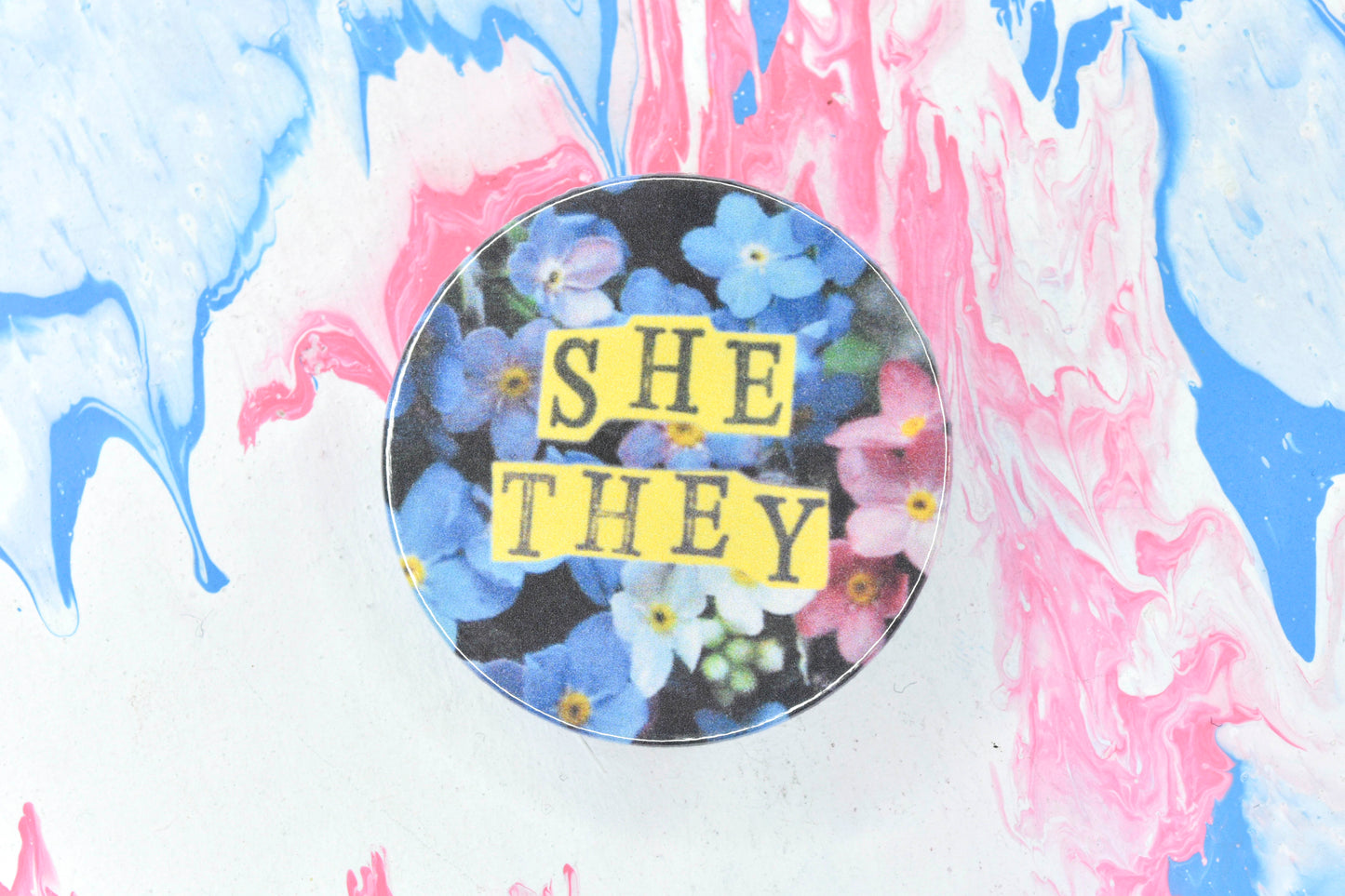 A pin badge featuring a floral background showing some pink, white and blue forget-me-not flowers. Overlaid are hand cut pieces of yellow paper with hand stamped uppercase black lettering reading "she" and "they".