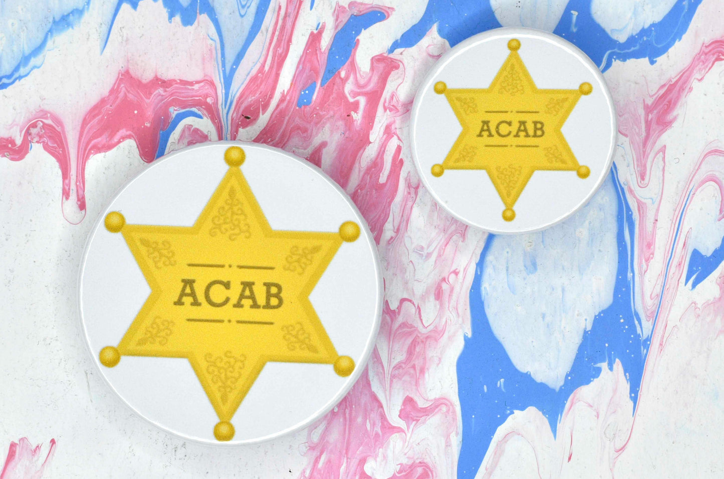 Two badges in sizes medium and small respectively, each featuring a white background with an illustration of a yellow/gold sheriff star. There is gold text on the star reading "ACAB"