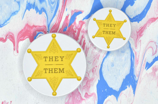 Two badges in sizes medium and small respectively, each featuring a white background with an illustration of a yellow/gold sheriff star. There is gold text on the star reading "they/them"