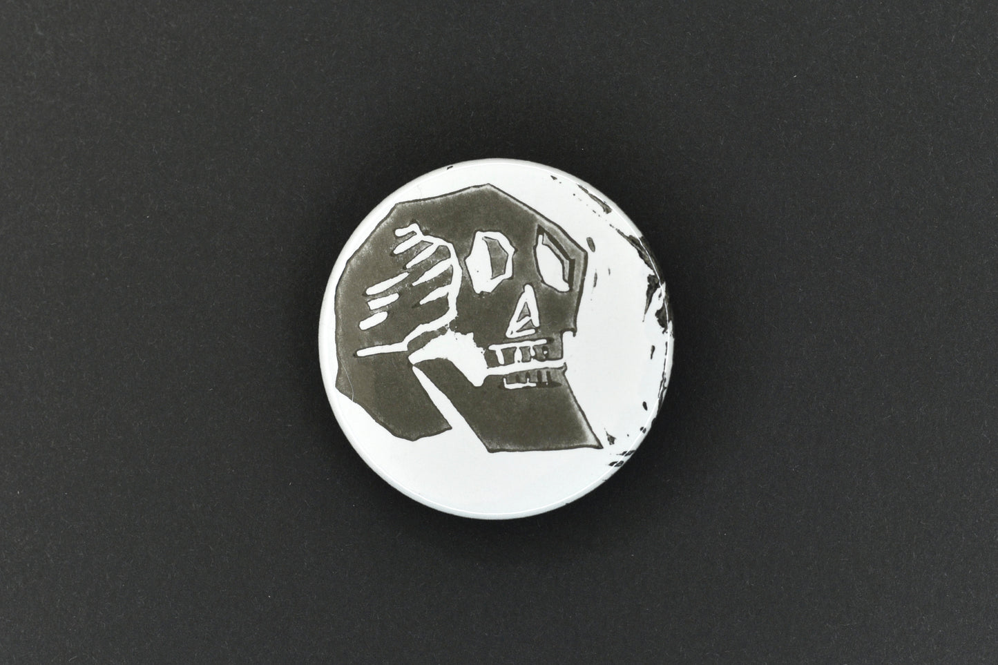 A single button badge featuring a white background with an original lino print stamped skull design in black ink.