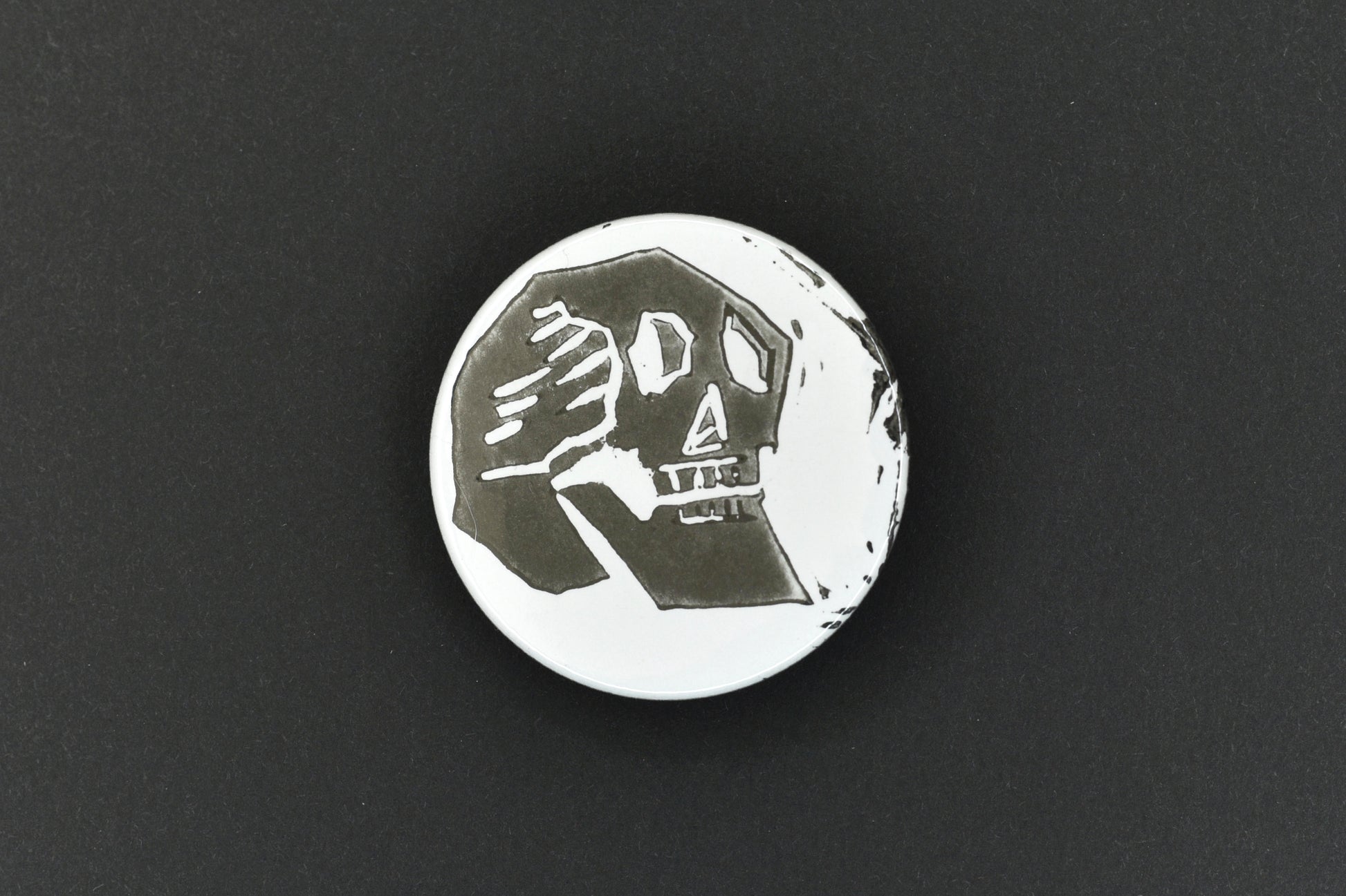 A single button badge featuring a white background with an original lino print stamped skull design in black ink.