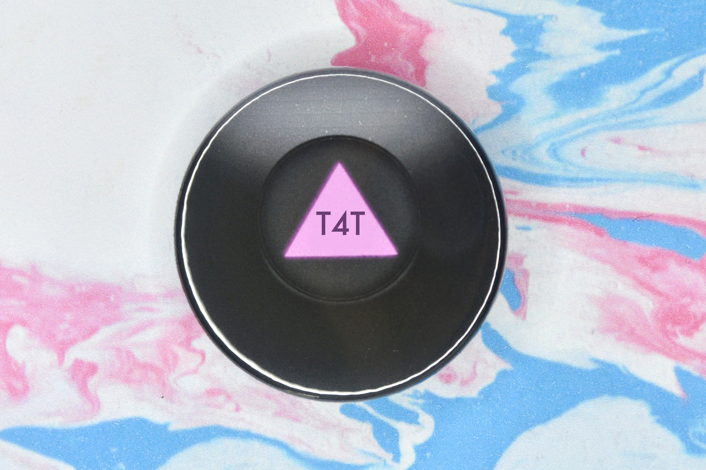 A circular badge in size medium featuring an illustration of a black magic 8 ball which takes up the full badge. There is a small circular window in the centre which shows a pink triangle with the message of the ball written on it in darker pink text. The text reads "T4T".