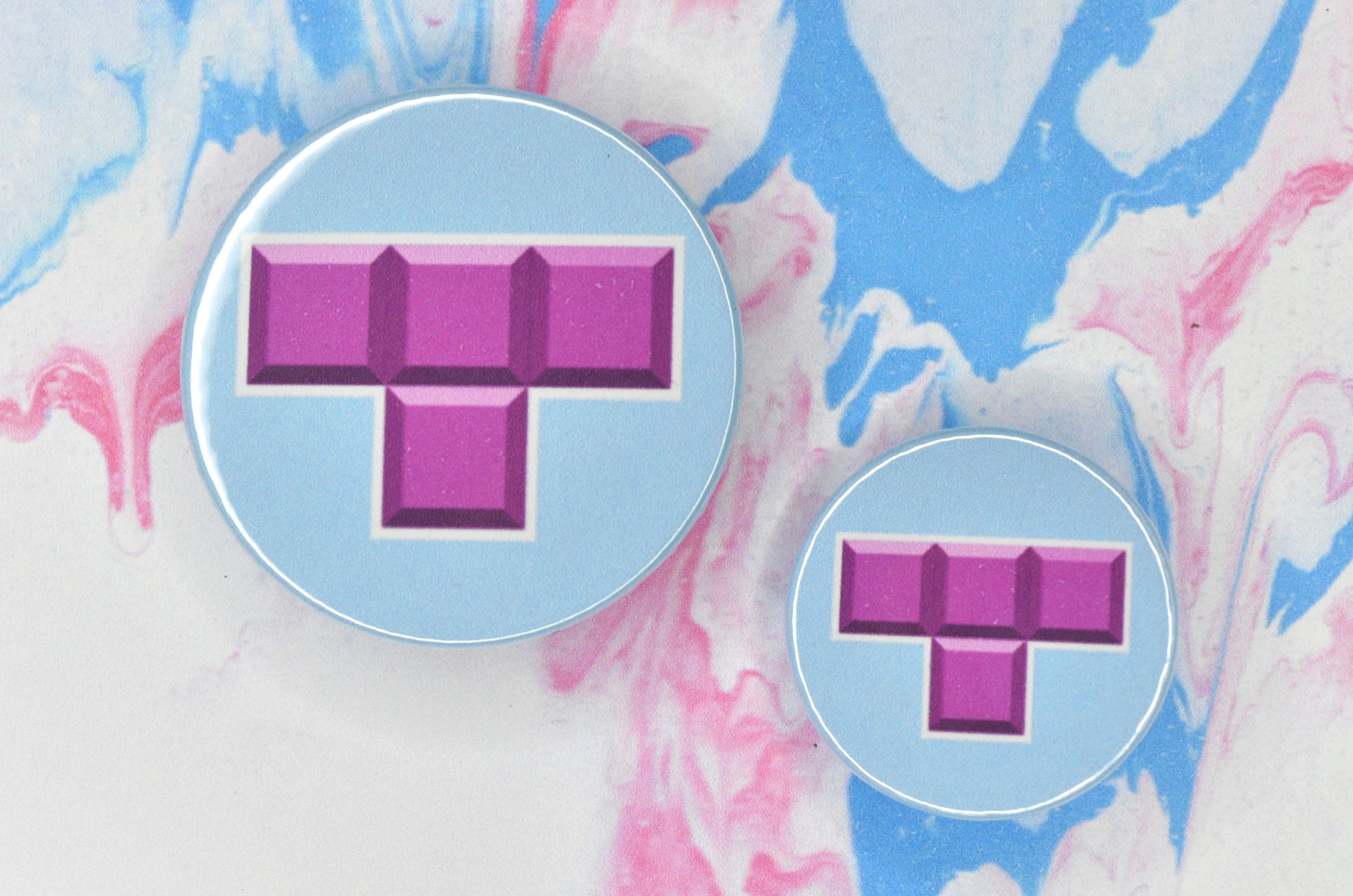 Two badges in sizes medium and small respectively, each featuring a light blue background with an illustration of a t-shaped Tetris block in pink with a slight white border around it.